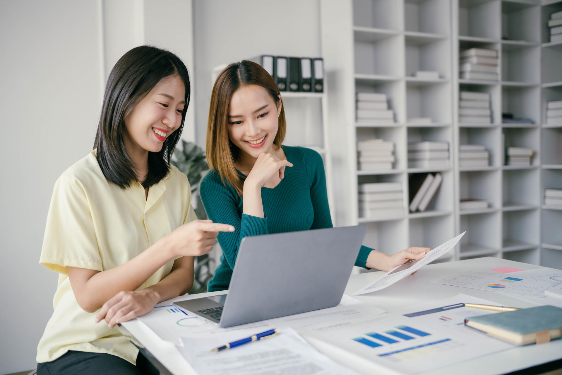 Two women are sitting at a desk with a laptop and papers. They are smiling and pointing at the laptop screen